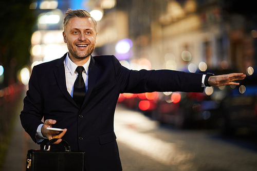 Happy man in formalwear outstretching his arm while catching taxi in urban environment