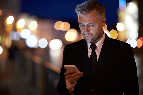 Serious man in formalwear texting in smartphone in urban environment at night