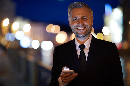Young cheerful businessman with smartphone looking at you with toothy smile outdoors