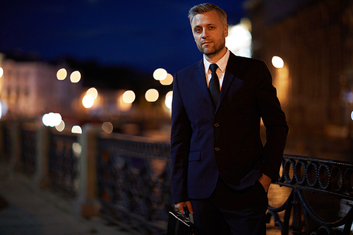 Elegant businessman in suit standing by riverside at night with lights on background