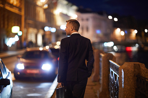Rear view of modern elegant businessman with briefcase walking along riverside at night