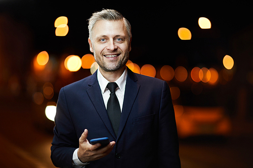 Young confident businessman with toothy smile messaging in smartphone in urban environment at night