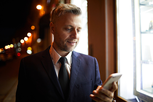 Businessman with smartphone standing by window of night shop while texting in urban environment