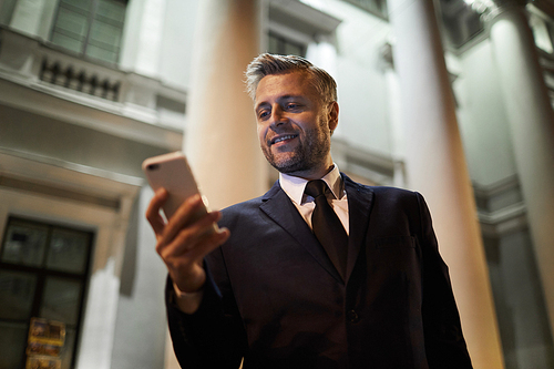 Contemporary mobile man in formalwear standing by modern building and messaging in smartphone at night