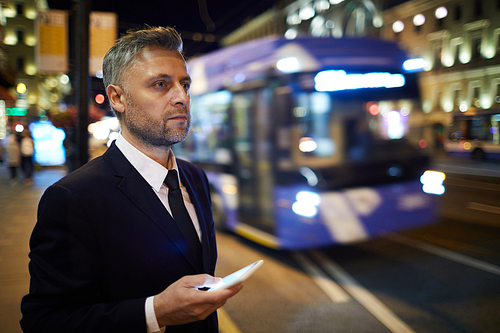 Serious businessman with smartphone ordering taxi cab while standing by road in the evening