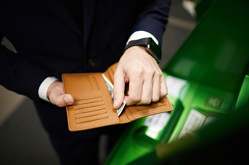 Hands of businessman putting banknotes into brown leather wallet after getting money from terminal