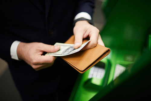 Businessman holding dollar banknote and leather wallet while standing by cash terminal