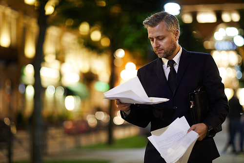 Busy young economist reading financial papers while moving along night street