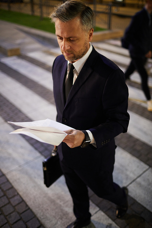 Busy young businessman in suit reading papers while crossing road at night