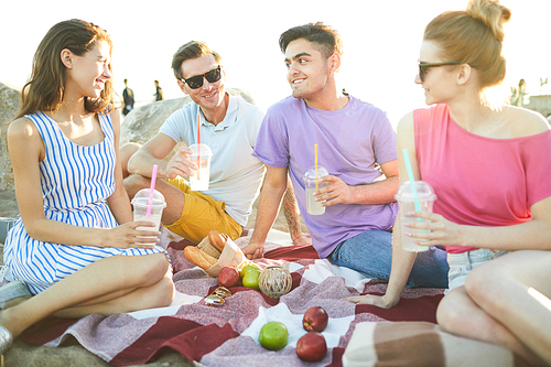 group of friendly guys and girls with drinks sitting on plaid in natural , talking and enjoying summer day