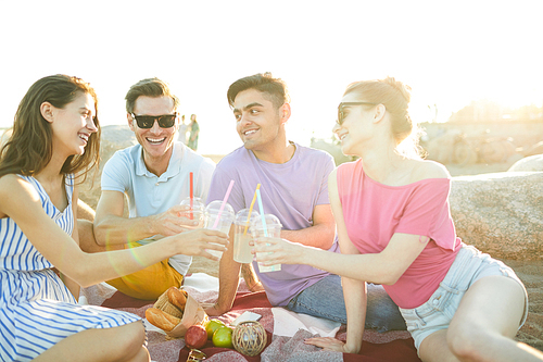 Four cheerful friends toasting with drinks while sitting on plaid and having picnic on sunny day