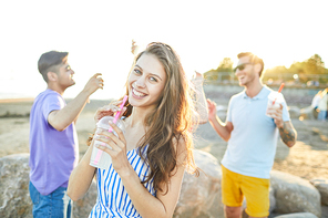 Cheerful girl with toothy smile having drink while her friends dancing on background on beach