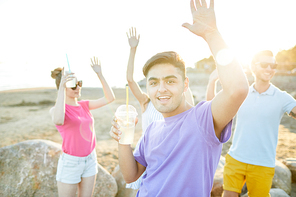 Excited guy with drink and his friends on background dancing on beach on summer day