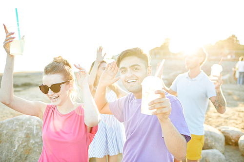 Ecstatic teenage friends with drinks having fun and dancing on the beach on sunny day