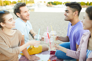 Two guys and their girlfriends with drinks toasting at picnic on the beach and talking