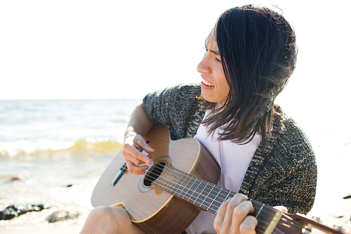 Handsome Asian man smiling and playing guitar while sitting on beach near sea.