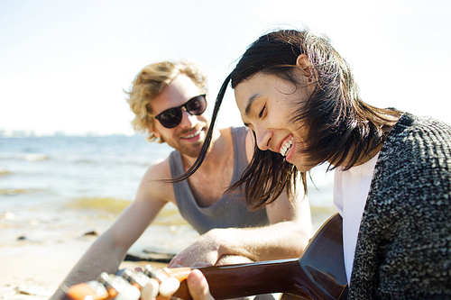 Two multiracial men smiling and playing guitar while spending time together on nice beach party.