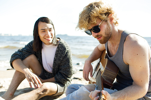 Two young men sitting on beach near sea and playing guitar during good party.
