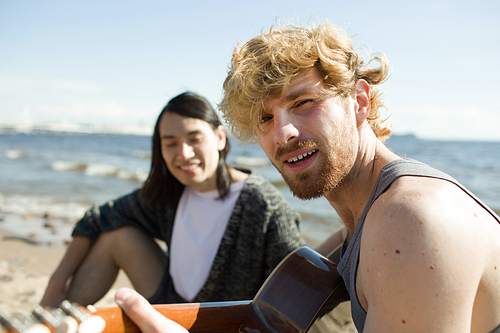 Attractive young man  and playing guitar while sitting on beach near Asian male friend.