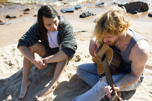 From above shot of two diverse men sitting on beach sand and playing guitar during party.
