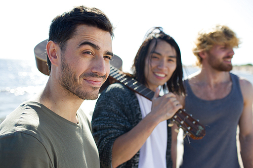 Three diverse friends with guitar standing on beach near sea.