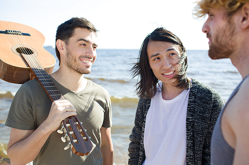 Three multiracial men with acoustic guitar smiling and communicating with each other while standing near sea together.