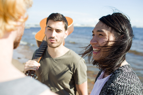 Three male friends with guitar smiling and talking with each other while standing on beach near sea.