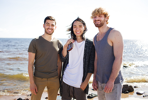 Three diverse young men with nice acoustic guitar smiling and  while standing on beach near sea.