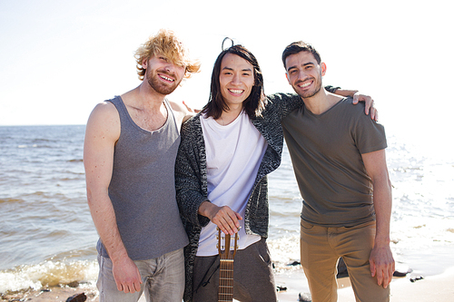 Three multiethnic men with acoustic guitar smiling and  while standing near sea.