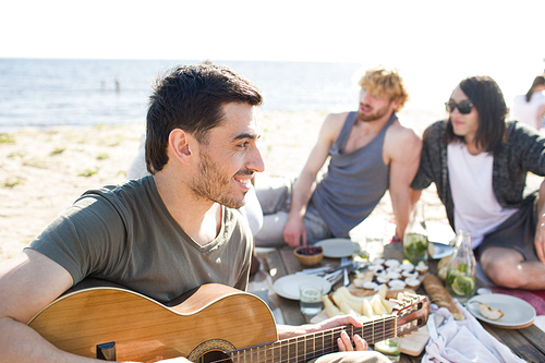 Attractive young man smiling and playing guitar while sitting on beach during multiethnic party.