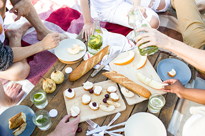 Group of crop people sitting around wooden board and eating tasty food during multiethnic party.