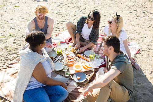 Group of diverse young people smiling and communicating with each other while having nice picnic during multiethnic party on beach.