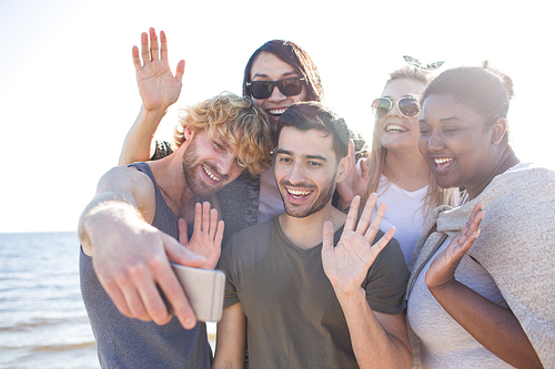 Group of diverse friends smiling and waving hands while standing on beach and posing for selfie during party.