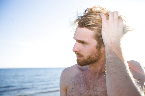 Attractive shirtless man touching hair and looking away while standing on beach near sea.