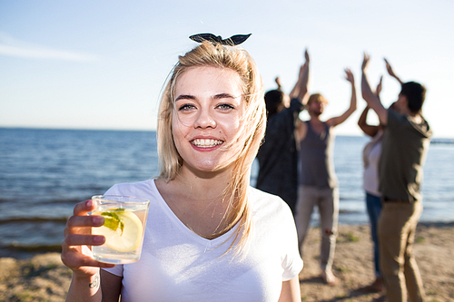 Beautiful young woman with glass of fresh beverage smiling and  while spending time on multiracial beach party.