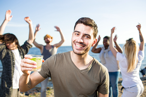 Attractive young man smiling and  while holding glass of fresh beverage during multiethnic party on beach.