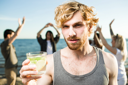 Handsome young man holding glass with cold drink and  while spending time on multiethnic party on beach.