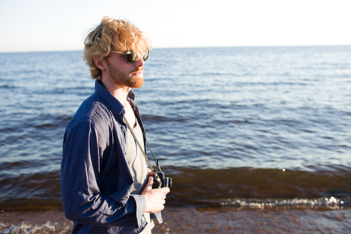 Handsome young man in casual outfit holding camera while walking on beach near waving sea.