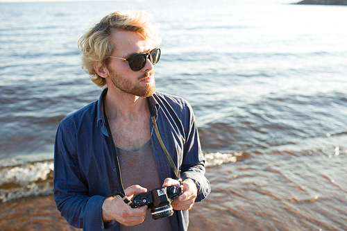 Attractive young man in sunglasses holding camera and looking away while standing on beach near waving sea.
