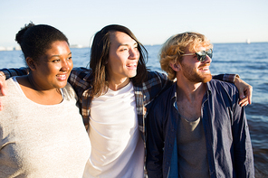 Group of diverse young people smiling and looking away while standing on beach near sea together.