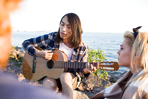 Handsome Asian man sitting on beach and playing guitar while spending time with friends on multiethnic party.
