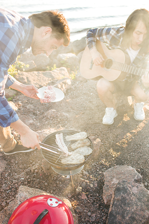 Two young men roasting meat on grill and playing guitar while spending time on multiethnic beach party.