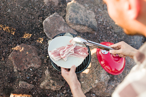 From above shot of faceless person using grill to fry delicious bacon and meat during party on beach.