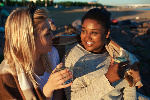 Two multiethnic young women smiling and looking at each other while sitting under blanket and drinking during beach party.