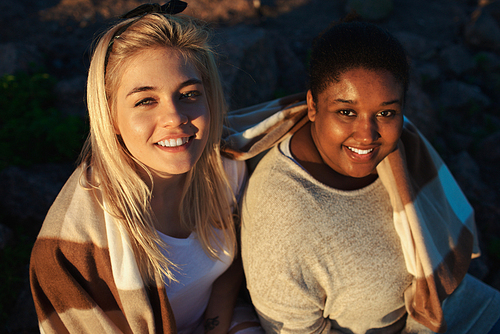 Two diverse young women smiling and  while sitting under blanket during good multiethnic party.