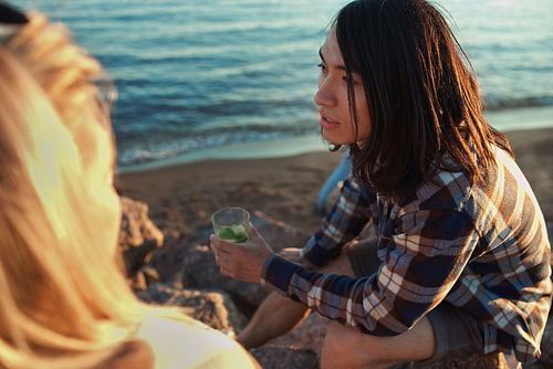 Handsome Asian man holding glass of cold beverage and talking with faceless woman while sitting on beach during multiracial party.