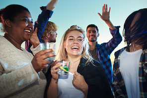 Lovely young woman holding glass of fresh beverage and laughing while standing amidst dancing multiracial friends during party.