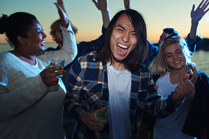 Cheerful Asian man with glass of cold drink having fun while standing amidst dancing friends on multiethnic party.