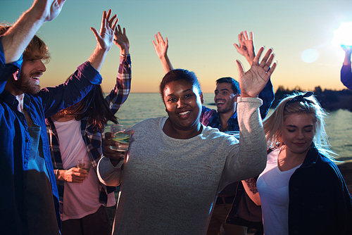 Young African-American woman with glass of cold beverage smiling and  while dancing among diverse friends on nice party.