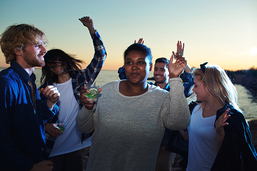 Young black woman holding glass of cold drink and dancing among friends while having fun during multiethnic party.